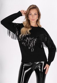 Fekete pulcsi hosszú rojttal és flitterekkel a vállnál, bordázott mandzsettával és aljjal. Fényes fekete leggings-szel párosítva.