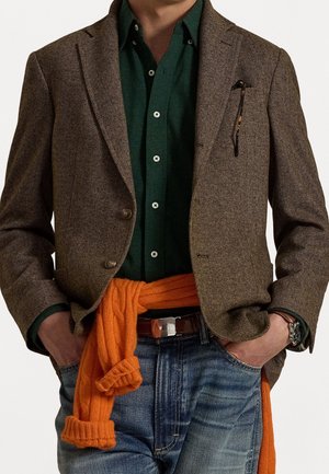 Mann trägt braunen Fischgrätblazer über dunkelgrünem Hemd, mit orangefarbenem Pullover um die Taille gebunden und blaue Jeans, Hände in den Taschen.