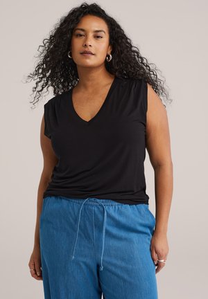 Frau mit lockigem Haar, die ein schwarzes ärmelloses V-Ausschnitt-Top und blaue Hose mit Kordelzug trägt, steht vor einem schlichten hellen Hintergrund.