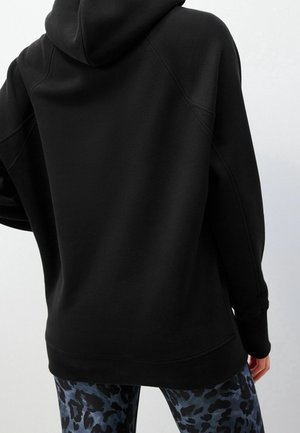 Sudadera negra de gran tamaño con una textura suave, que cuenta con capucha ajustable y puños acanalados. La tela tiene un acabado mate.