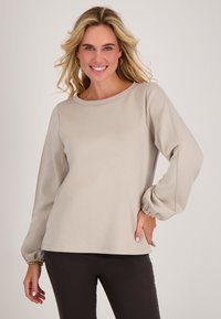 Beige longsleeve top met een ronde halslijn en gathered manchetten, gemaakt van zachte, gestructureerde stof. Gecombineerd met donkere, strakke broeken.