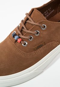 Baskets en daim marron dotées d'œillets argentés, de lacets marron et de trois bloque-lacets colorés (bleu, rouge, orange) avec une semelle en caoutchouc blanche.