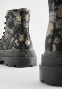 Bottes noires à motif floral comprenant un matériau texturé et matelassé, une semelle noire robuste et des accents métalliques sur le bord supérieur.