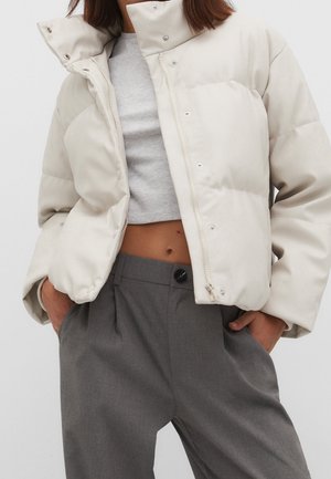 Mujer con chaqueta acolchada crema sobre un top gris corto y pantalones grises de cintura alta, con las manos en los bolsillos.