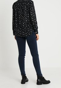 Blouse noire avec motif de feuilles blanches, design à boutons, associée à un jean skinny bleu foncé. Bottines noires avec semelles épaisses.