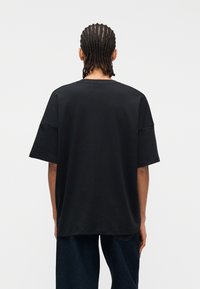 Άτομο με πλεξούδες, φορώντας φαρδιά μαύρη κοντομάνικη μπλούζα και σκούρο μπλε τζιν, στέκεται  απέναντι σε λευκό ομοιόμορφο φόντο.