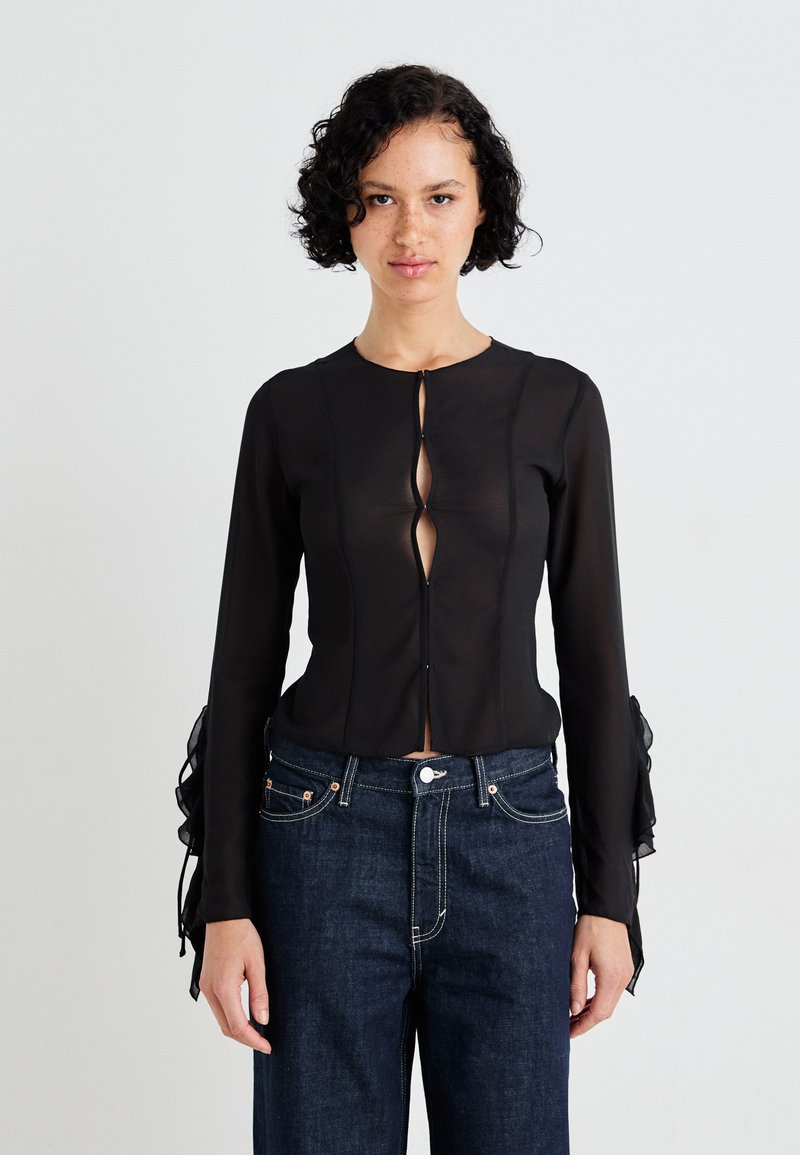 Top lung cu maneci lungi din material subțire negru, cu deschidere în formă de cheie în față și manșete volan. Purtați cu blugi negri din denim, cu detalii de cusătură vizibile.