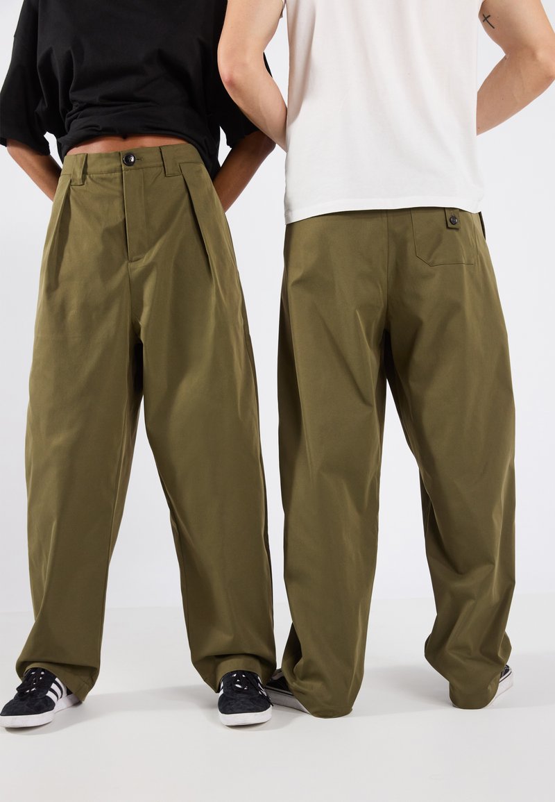 Pantalons larges vert olive avec plis, dotés d'une fermeture à bouton et d'une poche arrière. Le matériau est du coton avec une texture lisse.