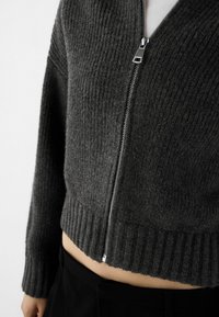 Pull cropped en maille côtelée grise avec une fermeture éclair à l'avant ; il présente un ourlet ajusté et des manches longues, mettant en valeur une texture douce.