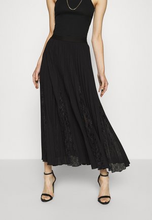 Femme portant un haut noir sans manches, une longue jupe noire plissée avec des inserts en dentelle, et des sandales à talons hauts noires, debout devant un fond clair.