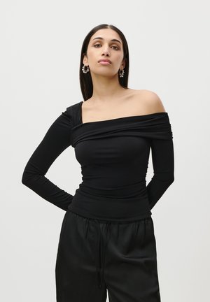 Black long-sleeve top with one shoulder exposed, featuring gathered draping and smooth texture. Paired with black, relaxed-fit pants.