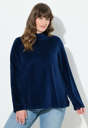 Top de terciopelo azul marino con cuello vuelto, que presenta mangas largas, un corte relajado y aberturas laterales para mayor libertad de movimiento. Textura suave, diseño minimalista.