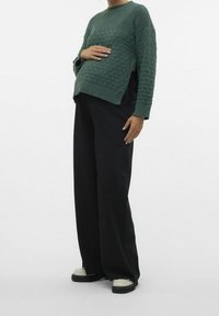 Zielony sweter dziany z teksturowanym wzorem w romby, o okrągłym dekolcie i długich rękawach, zestawiony z szerokimi czarnymi spodniami i casualowymi butami.