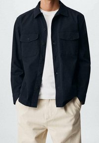 Granatowa koszula zapinana na guziki z dwoma kieszeniami na piersi, wykonana z gładkiego materiału. Nosi się ją na białym T-shircie i zestawia z jasnymi spodniami.