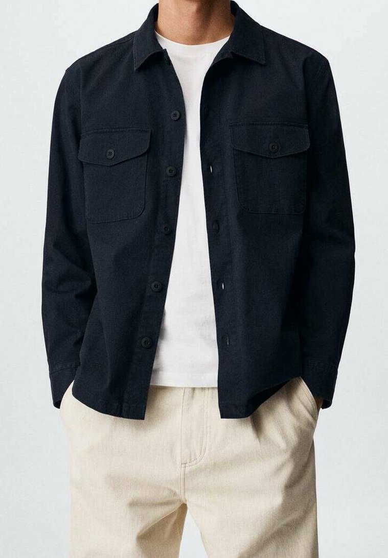 Granatowa koszula zapinana na guziki z dwoma kieszeniami na piersi, wykonana z gładkiego materiału. Nosi się ją na białym T-shircie i zestawia z jasnymi spodniami.