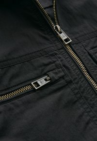 Veste noire avec une fermeture éclair dorée bien visible, texture de tissu lisse et détail de poche zippée, mettant en avant des éléments de design moderne.