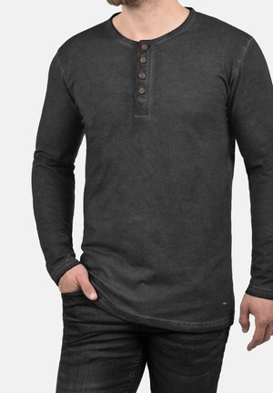 Schwarzes langärmliges Henley-Shirt aus weichem Stoff mit rundem Halsausschnitt, fünf braunen Knöpfen und subtiler Verblassungstextur. Kombiniert mit dunklen Jeans.