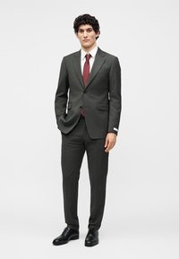 Άνδρας στέκεται φορώντας σκούρο γκρί κοστούμι με λευκό πουκάμισο, μπορντό γραβάτα και μαύρα επίσημα παπούτσια μπροστά σε απλό φωτεινό φόντο.