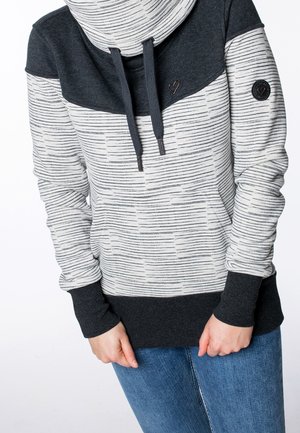 Personne portant un sweat à capuche rayé gris et noir avec des poignets noirs et une poche avant, associé à un jean bleu, mains jointes devant.
