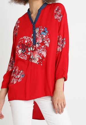 Rode blouse met lange mouwen met bloem-hartpatronen en blauwe vlinderdetails, blauwe halslijn, gedragen met witte broek.