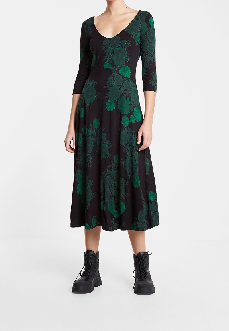 Robe noire mi-longue aux genoux avec imprimé floral vert, col en V et manches trois-quarts, associée à des bottes de combat noires à lacets.