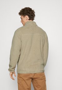 Beżowy polarowy sweter z wysokim kołnierzem, teksturowaną powierzchnią i długimi rękawami. Prosty fason bez widocznych logotypów czy wzorów.