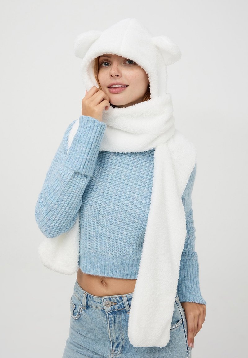 Biały puszysty szalik z przymocowanym kapturem, w którym znajdują się uszy misia, zestawiony z jasnoniebieskim swetrem z dzianiny. Miła w dotyku tekstura; swobodny design.