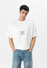 T-shirt en coton blanc avec une coupe ample, arborant un design graphique noir détaillé sur la poitrine. Assorti avec un jean en denim bleu clair.
