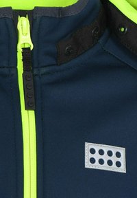Marineblauer Sakko mit einem leuchtend gelben Reißverschluss, schwarzen Akzenten und einem grauen Abzeichen mit sechs ausgeschnittenen Kreisen. Glatte Stoffstruktur.