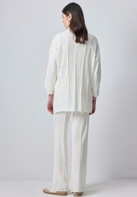 Túnica blanca de punto y pantalones a juego con textura de canalé vertical, que presentan un corte relajado y mangas largas. Zapatillas beige completan el conjunto.