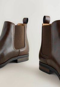 Botas Chelsea de cuero marrón oscuro, con paneles laterales elásticos y lengüetas, mostradas desde un ángulo sobre un fondo claro.