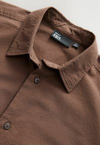 Camisa marrón hecha de un tejido texturizado, con un collar clásico, dos botones y una etiqueta con "NEXT EDIT."