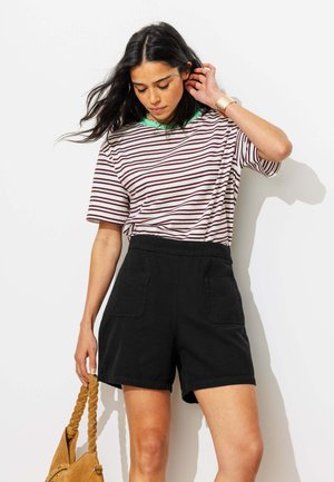 Femme portant un t-shirt rayé et un short noir, tenant un sac tressé beige, debout contre un mur blanc, touchant ses cheveux d'une main.