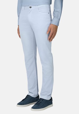 Mand iført lyseblå slim-fit bukser, matchende lyseblå skjorte og mørkeblå sneakers med hvide såler, stående mod en hvid baggrund.