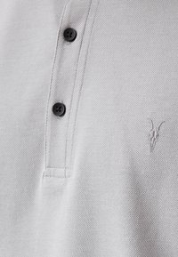 Grijze poloshirt van een gestructureerde stof, met twee zwarte knopen en een subtiel geborduurd logo aan de linkerkant.