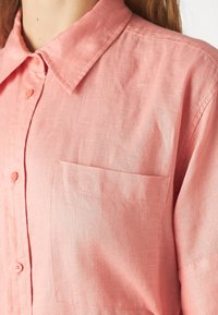 Camicia in lino rosa chiaro con colletto a punta, chiusura frontale con bottoni e una tasca applicata sul lato sinistro, che mostra un tessuto texturizzato.
