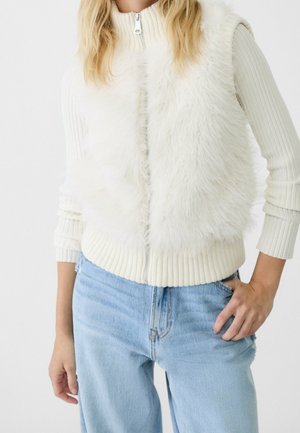 Kvinde iført en hvid, pelsagtig vest med lynlås over en ribbet cremefarvet sweater og lyseblå jeans, hånd hvilende i lommen.