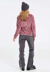 Pullover in pile di colore bordeaux con una finitura testurizzata, abbinato a pantaloni da sci grigi con cerniere laterali e scarponi da sci neri.