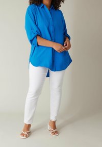 En person klädd i en lös blå skjorta med knappar, vita åtsittande byxor och vita sandaler med klack och remmar, som står mot en ljus bakgrund.