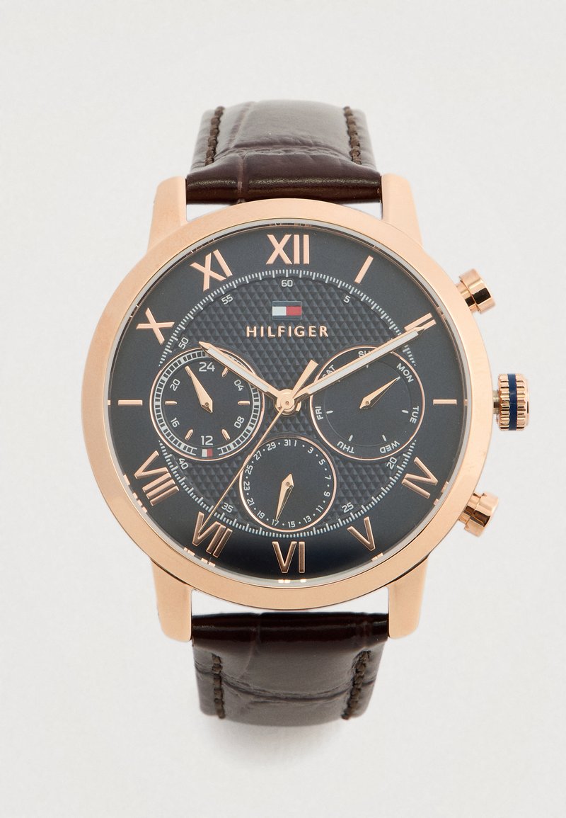 Orologio da polso rotondo con quadrante scuro, cassa in oro rosa, numeri romani, tre sottodial per 24 ore, giorno e data, e cinturino in pelle marrone.