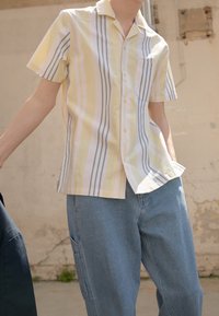 Camicia a maniche corte a righe in giallo pastello e blu tenue, con colletto, bottoni e taschino; indossata con pantaloni in denim blu chiaro.