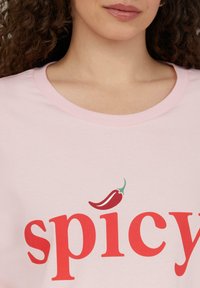 Lichtroze t-shirt met een vetgedrukt rode tekst die "spicy" zegt, met een kleine afbeelding van een rode chilipeper boven de letter 'i'.