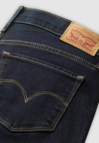 Jean en denim bleu foncé avec une étiquette de marque en cuir marron. Présente des coutures jaunes et un design de poche arrière courbée.