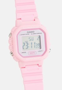 Montre numérique rose avec un boîtier carré, bracelet en caoutchouc, affichage LCD montrant l'heure, l'alarme et la résistance à l'eau. Comprend des boutons étiquetés MODE et LIGHT.