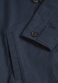 Svetlomodrá herringbone sako s detailmi gombíka a vrecka. Štruktúrovaná tkanina s hladkým povrchom a prispôsobeným vzhľadom.