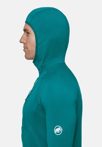 Mężczyzna w turkusowej kurtce z kapturem na zamek błyskawiczny z logo mamuta na rękawie, przedstawiony w profilu na jednolitym tle.