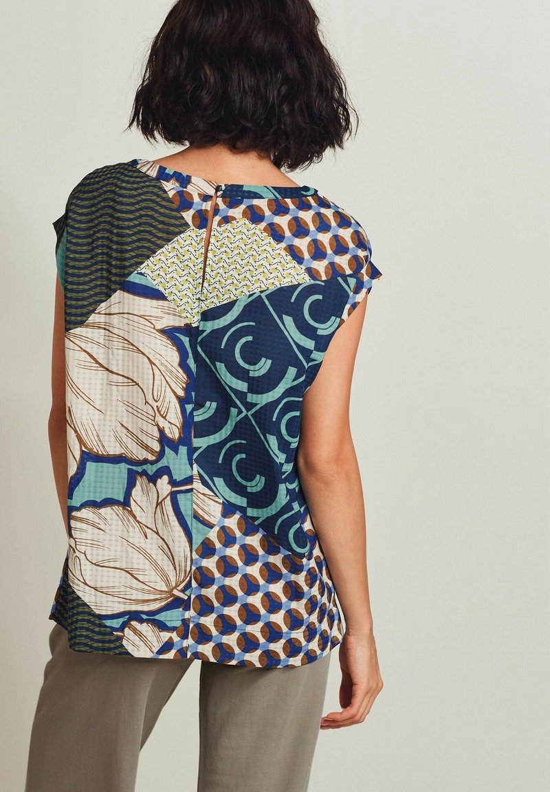 Femme portant un chemisier à manches courtes multicolore avec des motifs géométriques et floraux, associé à un pantalon gris clair, de dos.