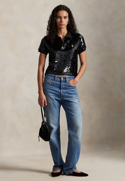 Črna polo majica z bleščicami in kratkimi rokavi, kombinirana s svetlo modrimi kavbojkami s pravimi hlačnicami. Manekenka drži majhno črno torbico.