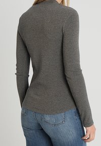 Graues Langarmoberteil mit figurnaher Passform, ausgestattet mit einem hohen Halsausschnitt und strukturiertem Stoff. Kombiniert mit blauen Jeans, die einen geschwungenen Saum zeigen.