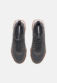 Graue Sportschuhe mit perforierten Details, strukturiertem, synthetischem Obermaterial, dunklen Schnürsenkeln und brauner Gummisohle. Timberland-Logo auf der Zunge.
